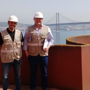 Fotografia de dois colaboradores do CRPG, nas instalações da SOVENA, de colete e capacete, com a ponte 25 de abril como pano de fundo