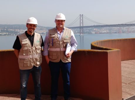 Fotografia de dois colaboradores do CRPG, nas instalações da SOVENA, de colete e capacete, com a ponte 25 de abril como pano de fundo