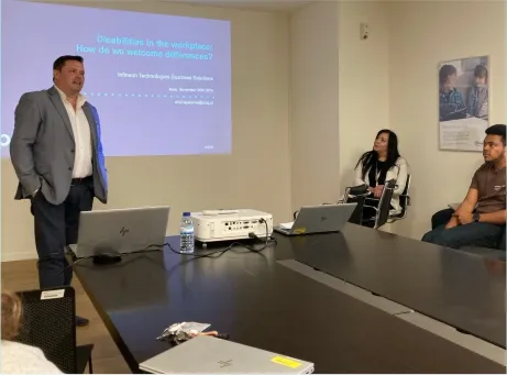 pessoa em pé, de mãos nos bolsos a falar com apresentação na parede e pessoas sentadas a assistir