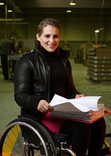 mulher,que se desloca em cadeira de rodas,com capa de folhas sobre as pernas em ambiente industrial