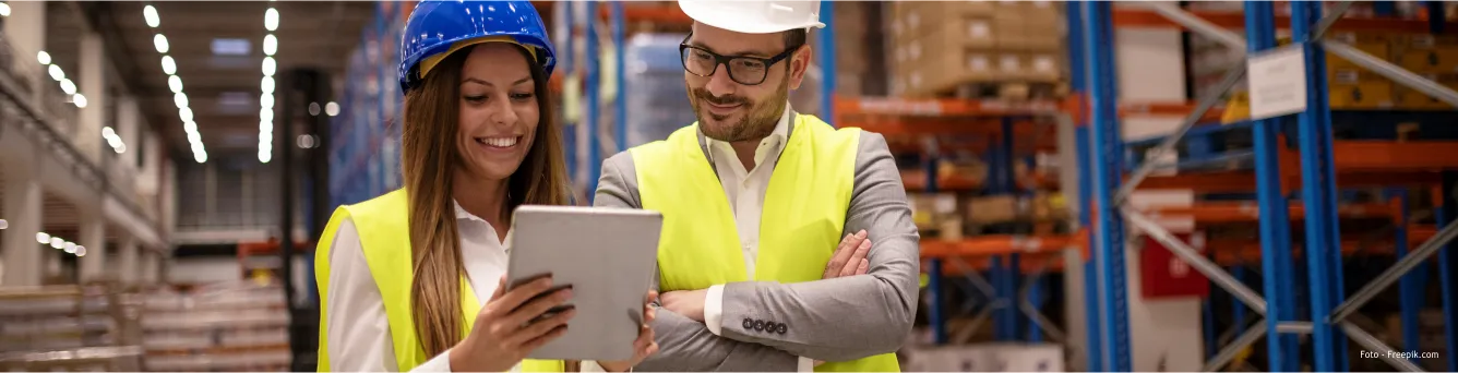imagem de duas pessoas, num armazém, com coletes refletores vestidos e capacete de proteção na cabeça enquanto olham para um tablet