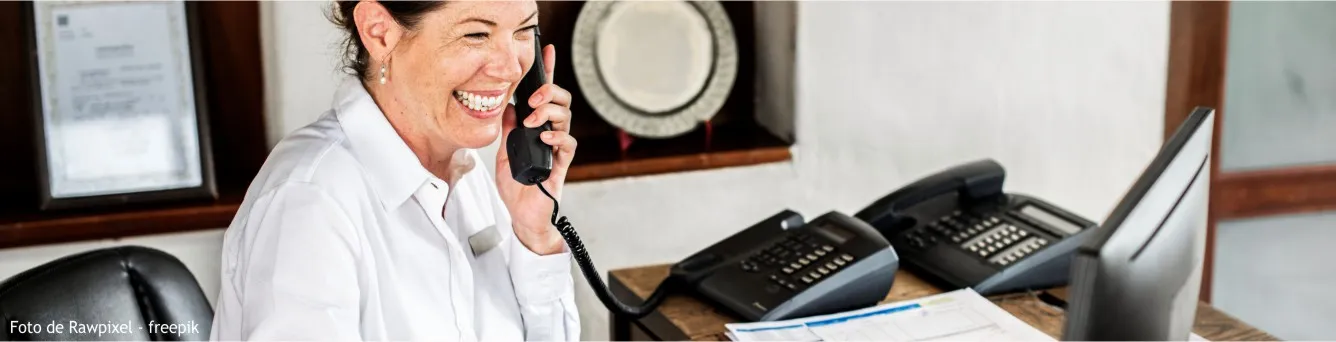 Mulher a falar ao telefone, à secretária, diante de um computador.