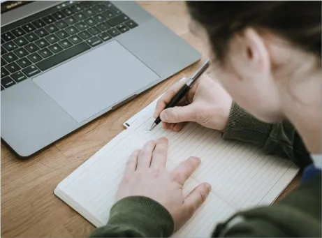 Mulher a escrever em caderno, com esferográfica, em frente a computador portátil