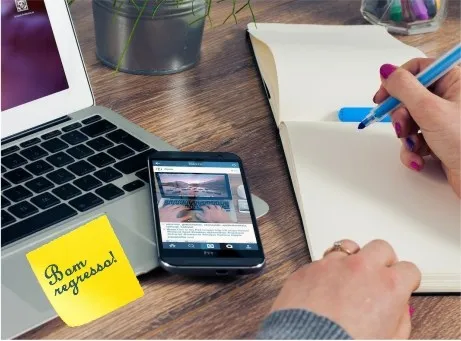 Mulher com caneta de feltro e caderno liso com telemóvel aberto num post de instagram. computador portátil e post-it com inscrição: 