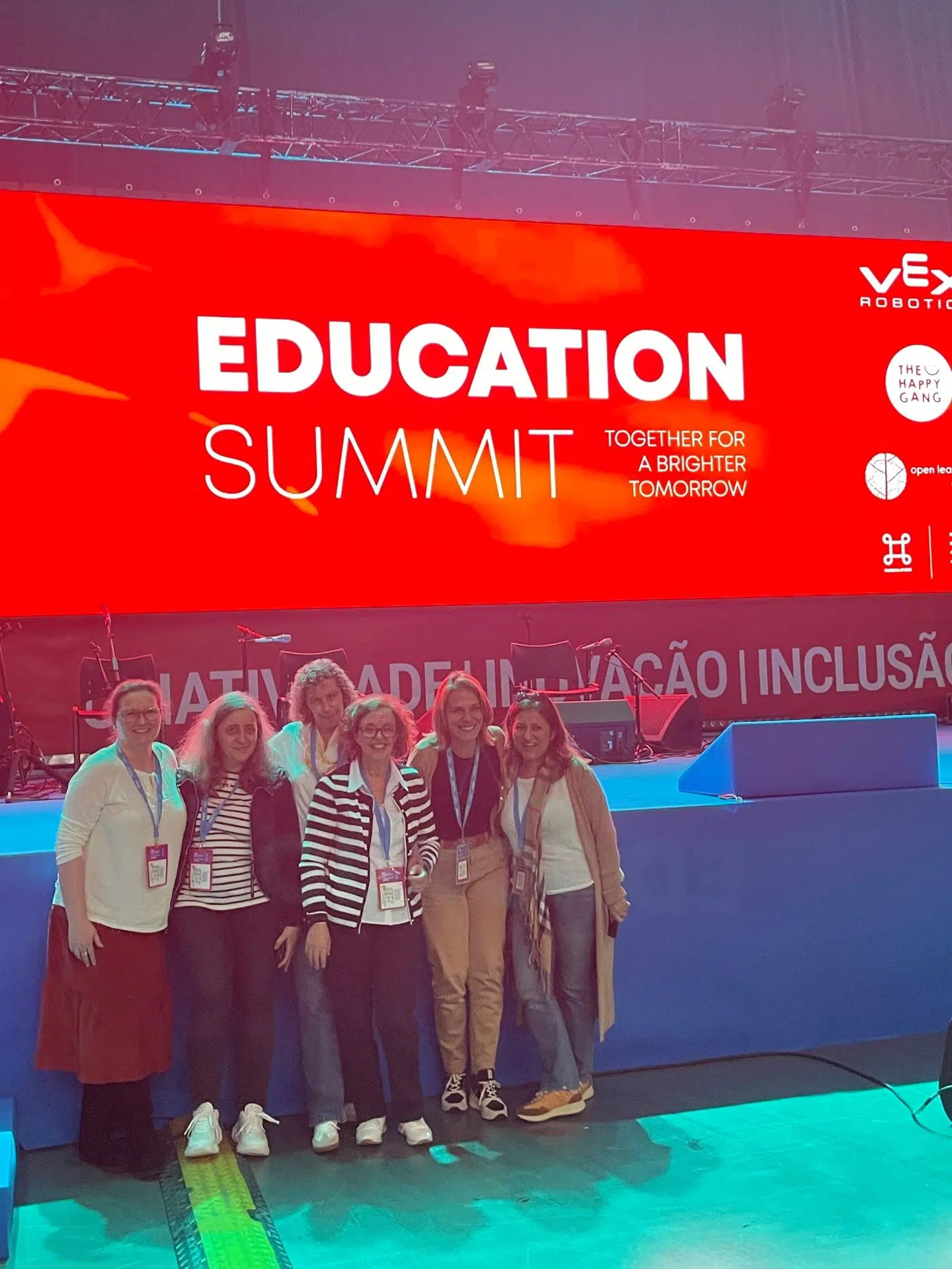 EducationSummit fotografia de 6 pessoas a sorrir para a camera, com palco nas suas costas e uma projeção na tela indicando Education Summit