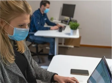 Homem e Mulher, ambos com máscara, sentados em secretárias, frente a computadores portáteis