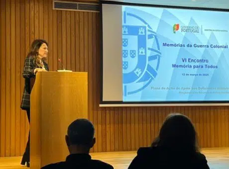 Fotografia de pessoa no palco, atrás de um pulpito a fazer uma apresentação e pessoas na plateia a assistir.