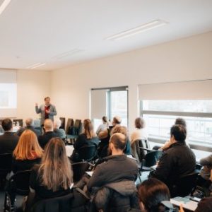 fotografia de pessoas sentadas a assistir a apresentação projetada na parede e orador em pé