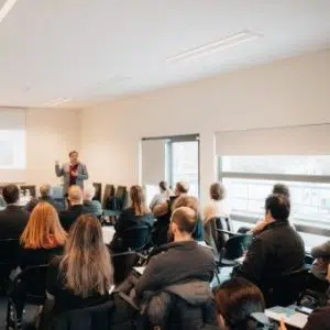 fotografia de pessoas sentadas a assistir a apresentação projetada na parede e orador em pé
