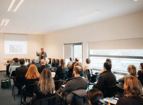 fotografia de pessoas sentadas a assistir a apresentação projetada na parede e orador em pé