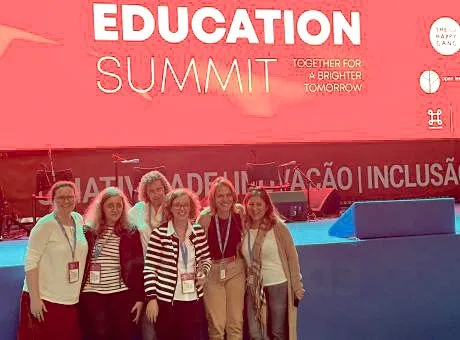fotografia de 6 pessoas a sorrir para a camera, com palco nas suas costas e uma projeção na tela indicando Education Summit