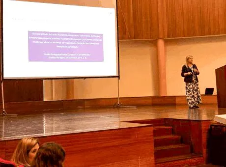 Fotografia de 1 pessoa em pé, num palco, a realizar uma apresentação. Apresentação a ser projetada na tela