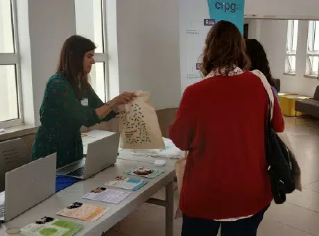 Fotografia do Ana Oliveira a sorrir, para duas pessoas que visitam o espaço do CRPG no evento. Roll-up do CRPG e duas mesas com folhetos