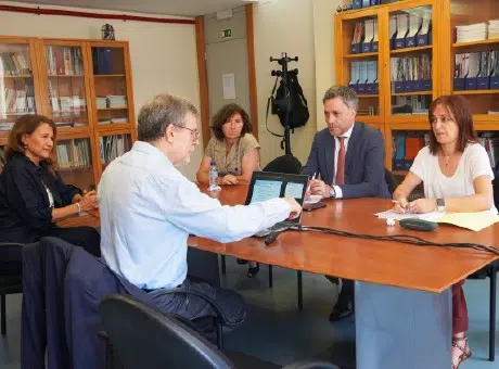 5 pessoas sentadas, a conversar, à volta de uma mesa, durante uma reunião. Computador e papeis em cima da mesa